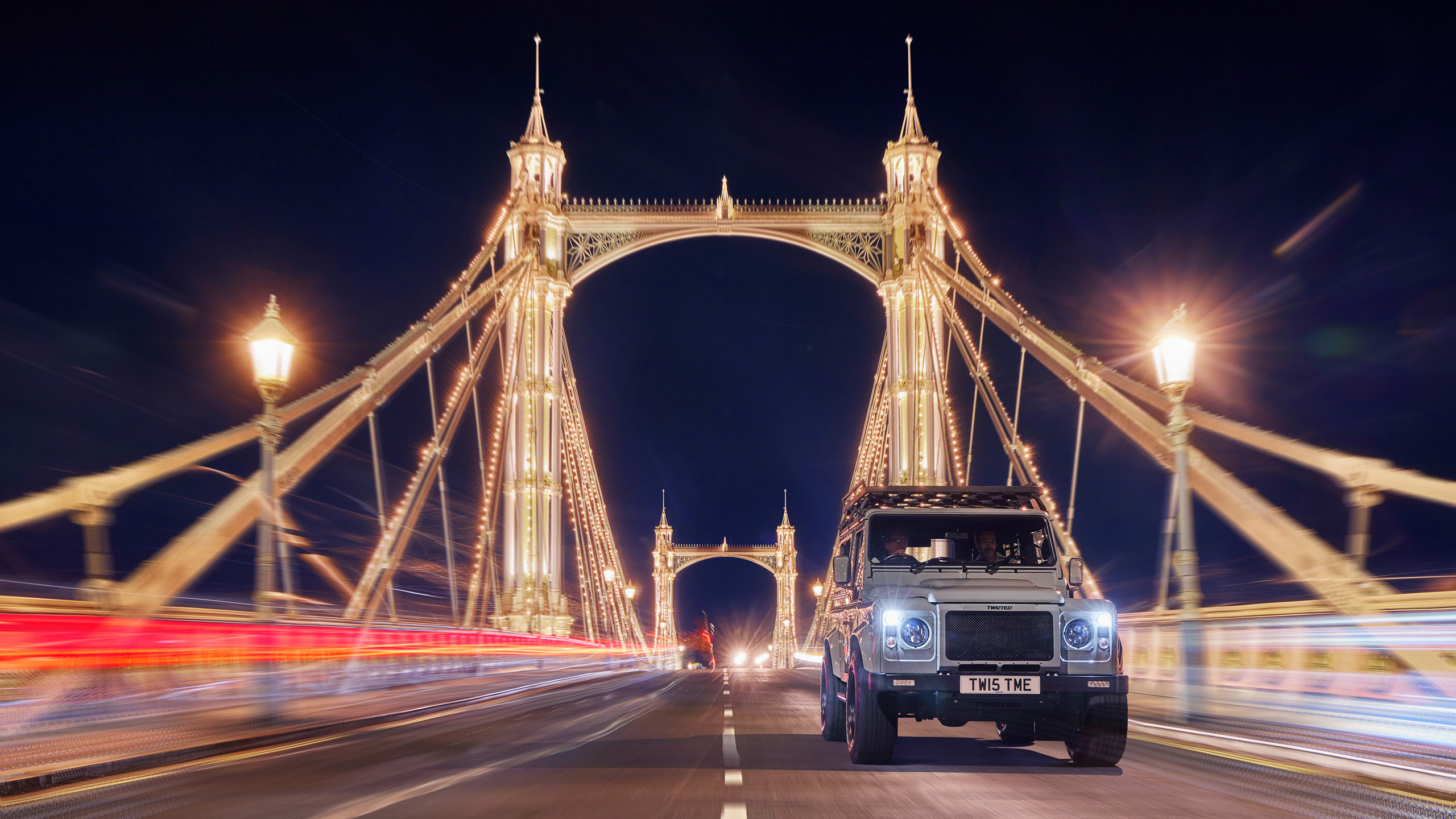 twisted-land-rover-defender-albert-bridge-night A Twisted Land Rover Defender driving across Albert Bridge in London at night, the bridge's lights illuminated.