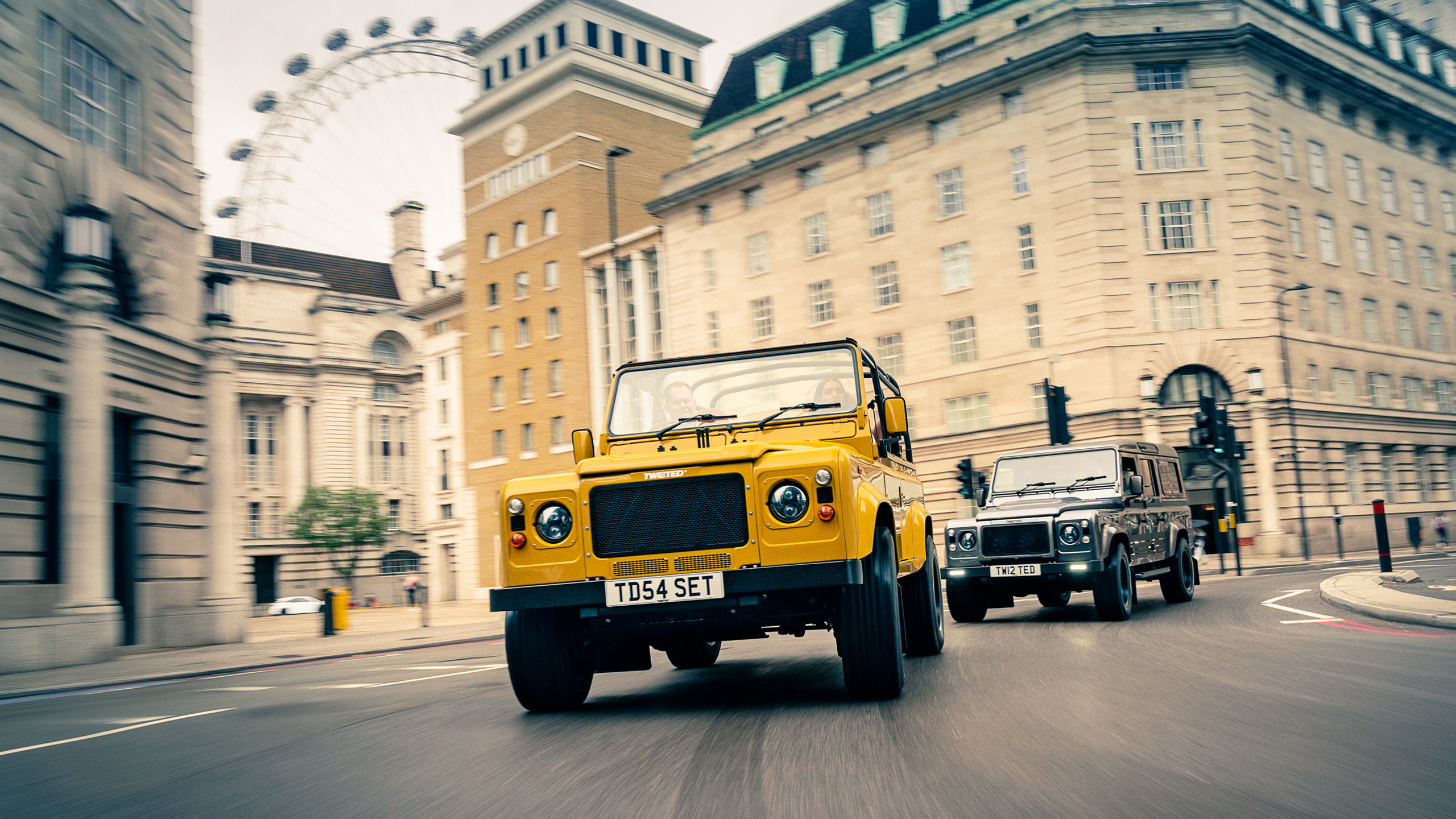 twisted-land-rover-defender-convoy-london-eye Two Twisted Land Rover Defenders, one yellow and one grey, driving through London with the London Eye visible in the background.